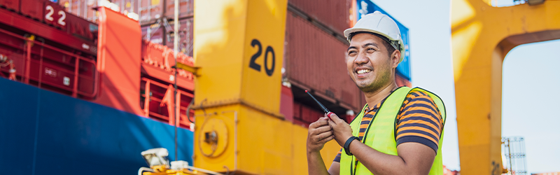portrait photo of happy logistics worker with radio transceiver working in port shipping containers.